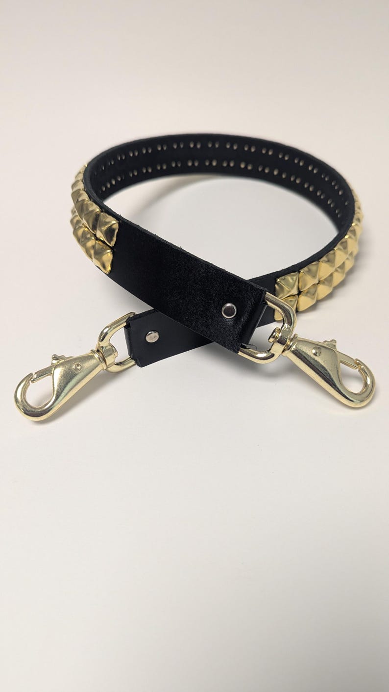 May include: Black handbag strap with gold-colored pyramid studs and gold-tone metal clasps. The strap is made of black leather and has a series of small holes along the top edge. The clasps are attached to the strap with metal rivets.