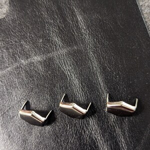 May include: Three silver-colored metal decorative rivets with a unique angular design. The rivets are arranged on a textured black leather surface, providing a contrast in color and texture. These rivets are likely used for leathercraft or adding embellishments.