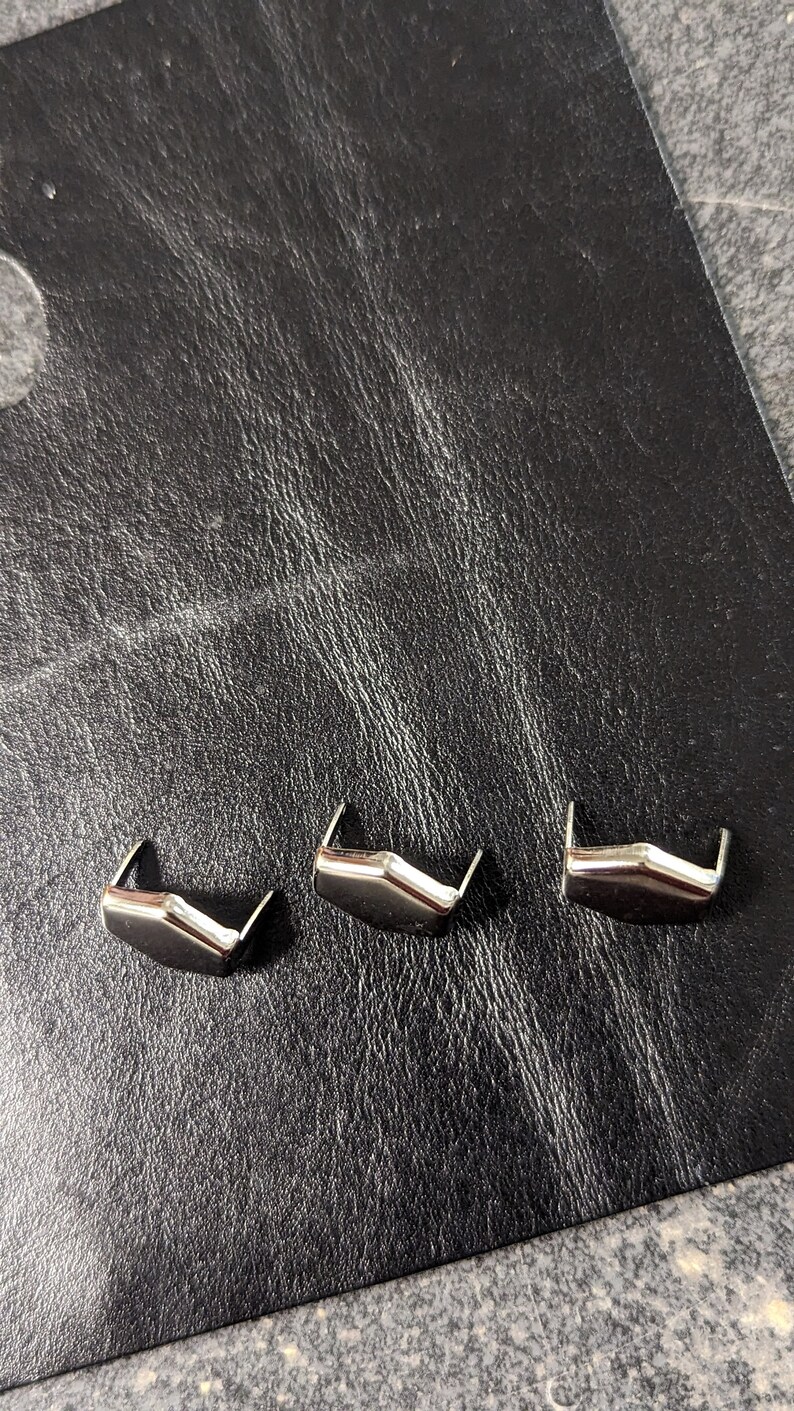 May include: Three silver-colored metal decorative rivets with a unique angular design. The rivets are arranged on a textured black leather surface, providing a contrast in color and texture. These rivets are likely used for leathercraft or adding embellishments.