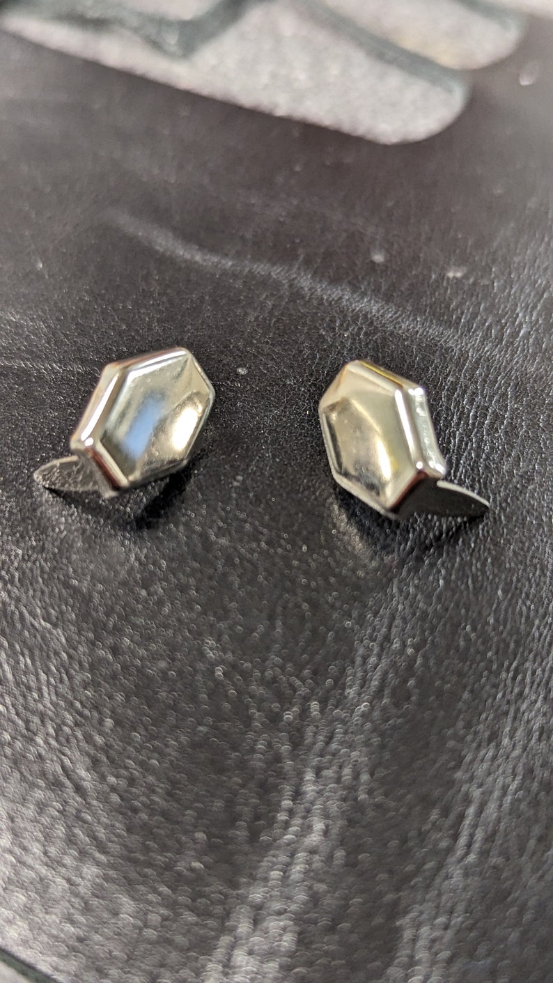May include: A pair of silver-toned metal stud earrings, each featuring a smooth, polished, hexagonal-shaped face with beveled edges. The earrings have a reflective surface and are designed with a post backing for secure attachment to the earlobe.