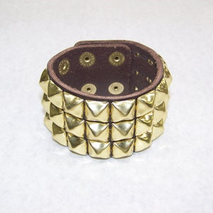 May include: A brown leather bracelet with three rows of gold pyramid-shaped studs.