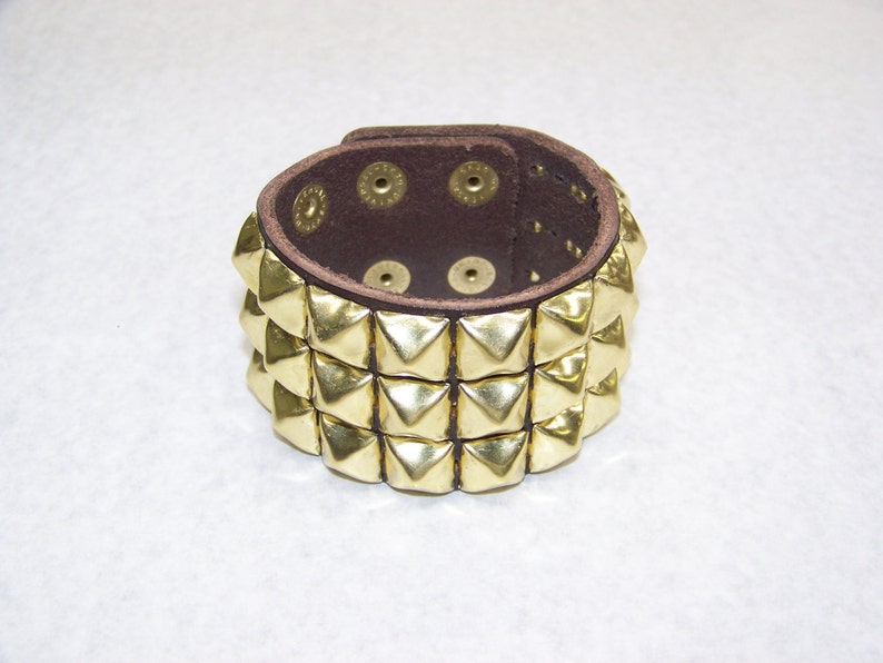 May include: A brown leather bracelet with three rows of gold pyramid-shaped studs.