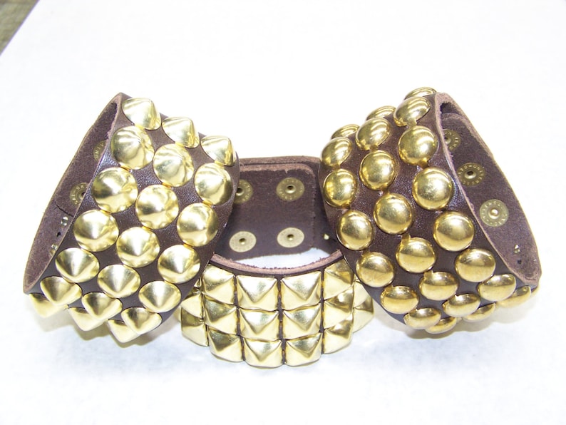 May include: Three brown leather bracelets with gold studs. The bracelets have different patterns of studs, including round, pyramid, and square shapes.