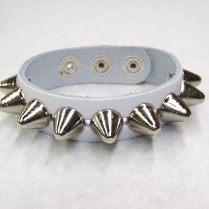 May include: White leather bracelet with silver pyramid-shaped studs. The bracelet has three snap closures.
