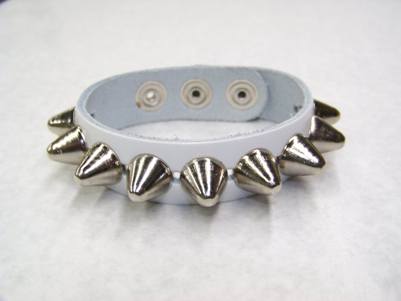 May include: White leather bracelet with silver pyramid-shaped studs. The bracelet has three snap closures.