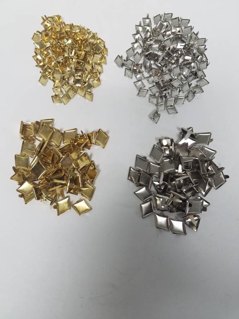 May include: Four piles of diamond-shaped metal studs in gold and silver tones. The studs are small, with a flat surface and a post on the back for attachment. The studs are scattered on a white surface.