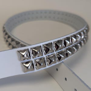 May include: White belt with two rows of silver square studs. The belt is made of a smooth, light-colored material and has several holes for adjustment. The studs are evenly spaced and reflect light.