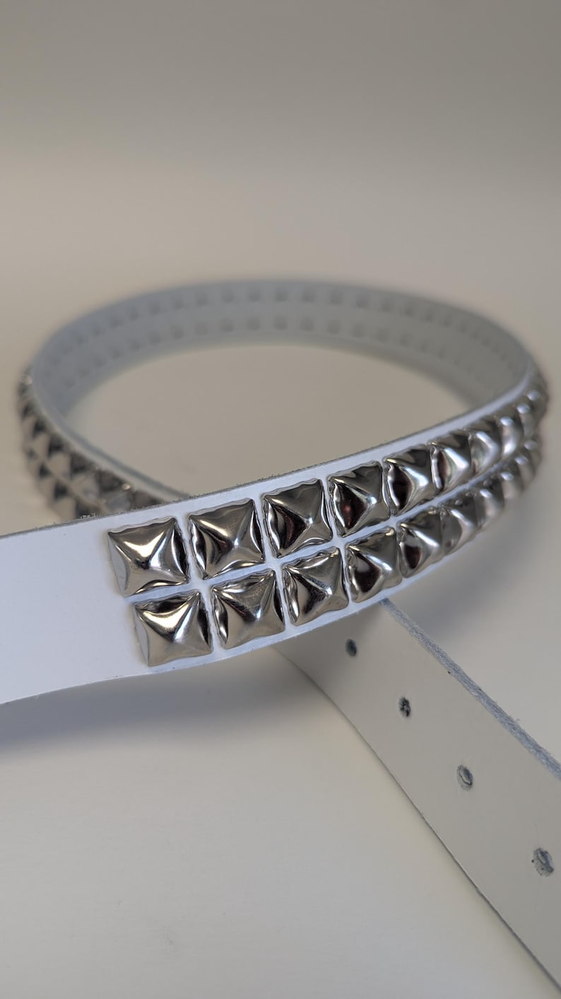 May include: White belt with two rows of silver square studs. The belt is made of a smooth, light-colored material and has several holes for adjustment. The studs are evenly spaced and reflect light.