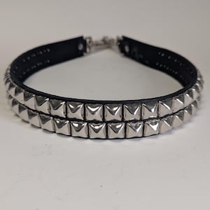 May include: A black leather choker necklace with two rows of silver-colored pyramid studs. The necklace has a silver-tone clasp and is displayed against a white background. This accessory is a bold statement piece.