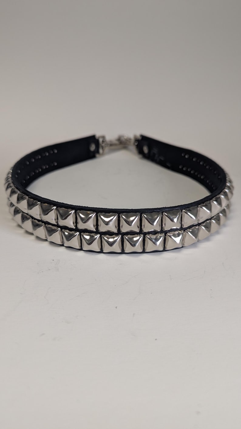 May include: A black leather choker necklace with two rows of silver-colored pyramid studs. The necklace has a silver-tone clasp and is displayed against a white background. This accessory is a bold statement piece.