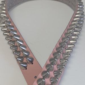 May include: A light pink belt with rows of silver-colored cone-shaped studs. The belt is made of a smooth material and has a buckle closure. The studs are evenly spaced along the length of the belt, creating a bold, edgy look.