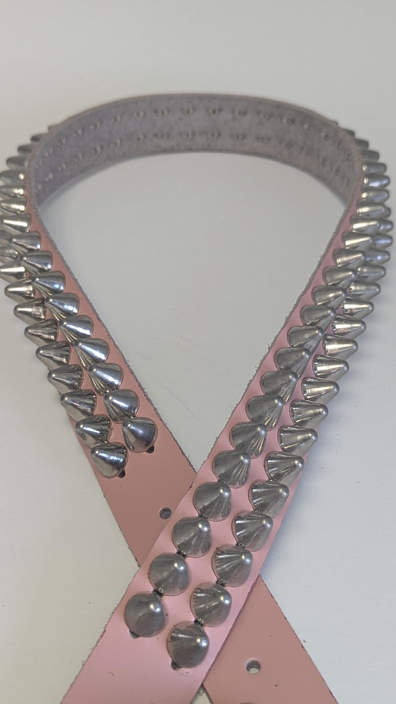 May include: A light pink belt with rows of silver-colored cone-shaped studs. The belt is made of a smooth material and has a buckle closure. The studs are evenly spaced along the length of the belt, creating a bold, edgy look.