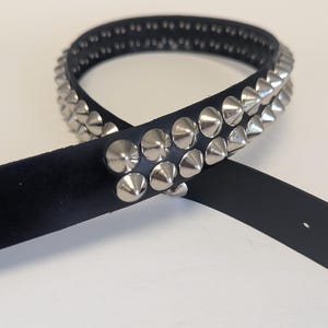 May include: Black leather belt with two rows of silver cone studs. The belt is shown on a white background. The belt has a single belt loop and a silver buckle. The belt is a fashion accessory.