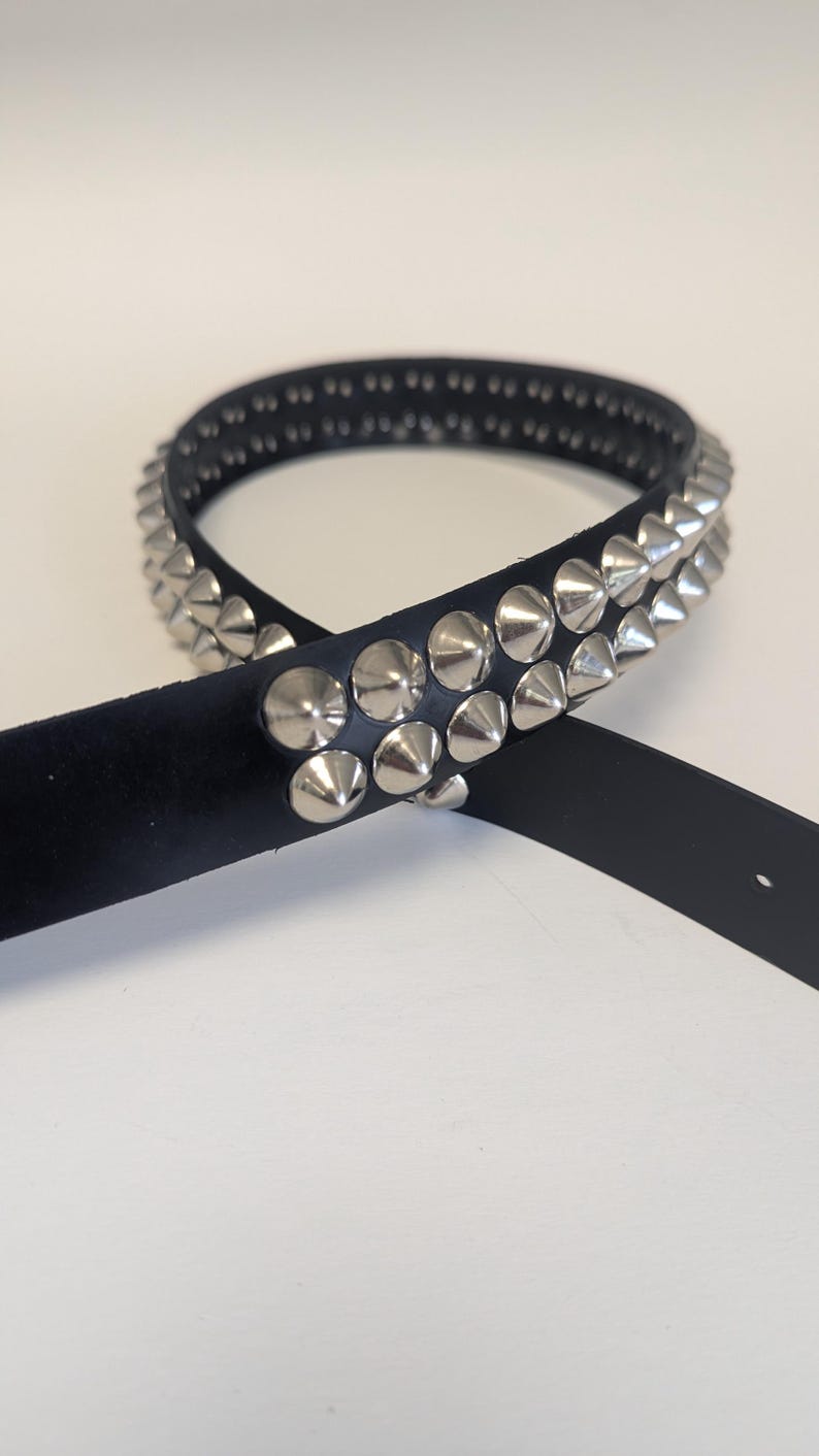 May include: Black leather belt with two rows of silver cone studs. The belt is shown on a white background. The belt has a single belt loop and a silver buckle. The belt is a fashion accessory.