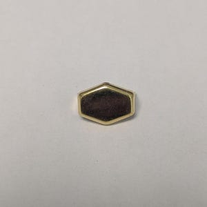 May include: A small, gold-colored, hexagonal object with a dark brown center. The object has a polished, reflective surface and a simple, geometric design. It is likely a decorative element or a small piece of jewelry.