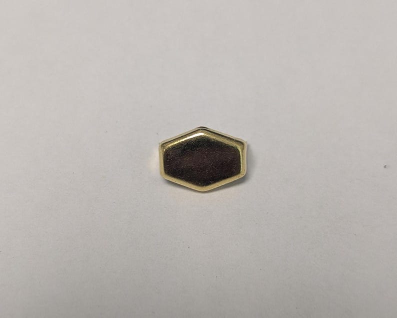 May include: A small, gold-colored, hexagonal object with a dark brown center. The object has a polished, reflective surface and a simple, geometric design. It is likely a decorative element or a small piece of jewelry.