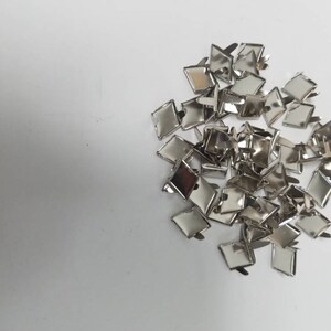May include: A collection of small, shiny, silver-colored metal findings, possibly for jewelry or crafts. Each piece has a square or diamond-shaped top with a clasp or pin on the back. The findings are scattered on a white surface.