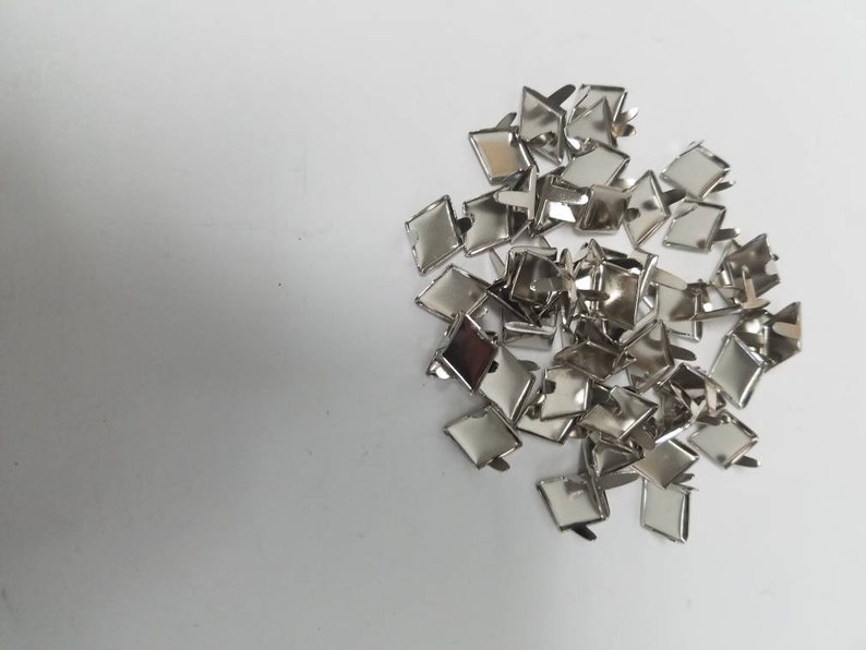 May include: A collection of small, shiny, silver-colored metal findings, possibly for jewelry or crafts. Each piece has a square or diamond-shaped top with a clasp or pin on the back. The findings are scattered on a white surface.