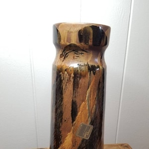 May include: Tall, cylindrical ceramic vase with a wide neck. The vase features a brown, black, and tan abstract pattern. A small tag that says "Pottery Craft" is attached to the side. The vase is displayed on a wooden surface.