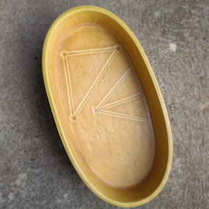 May include: A yellow oval-shaped ceramic dish with a geometric design in the center. The dish has a raised rim and a textured surface. The design consists of lines and triangles. The dish is likely for serving or display.