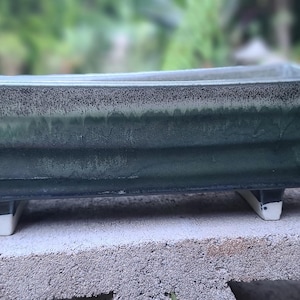 May include: A rectangular bonsai pot with a dark green glaze and a speckled rim. The pot is supported by four small, white-footed legs. The pot is sitting on a concrete block, with a blurred green background.
