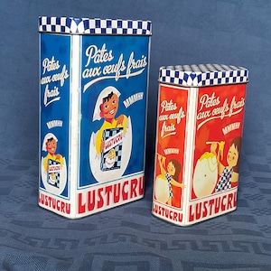 Peut inclure: Deux boîtes en étain rectangulaires vintage. La plus grande est bleue, la plus petite est rouge. Les deux ont un motif à carreaux sur les couvercles et présentent des illustrations d'enfants et d'œufs, avec du texte français.