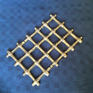 May include: Silver-coloured metal trivet with a lattice design. The trivet is made of interconnected bamboo-like rods, forming a diamond pattern. It sits on a blue fabric background.