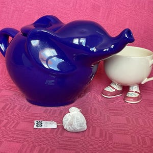 Peut inclure: Une théière en céramique bleue en forme d'éléphant avec une tasse à thé en céramique blanche avec des pieds roses. Un sachet de thé est au premier plan.