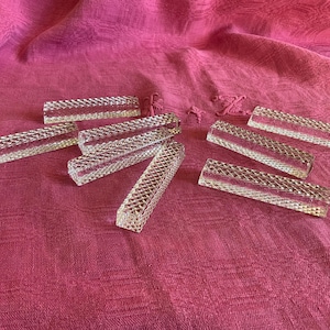 May include: Eight clear glass rectangular place card holders with a textured diamond pattern. The holders are arranged on a pink fabric surface.