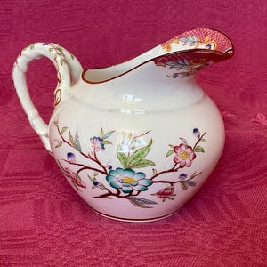 Può includere: Una brocca in ceramica bianca con un motivo floreale. La brocca presenta un manico curvo e un beccuccio con bordo decorato. Il motivo floreale include fiori e foglie rosa, blu e verdi.
