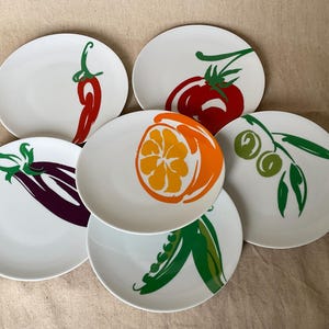 May include: A collection of six white ceramic plates, each decorated with a vibrant vegetable illustration. Designs include a red chilli, tomato, orange slice, aubergine, olives, and pea pods. Perfect for serving starters.