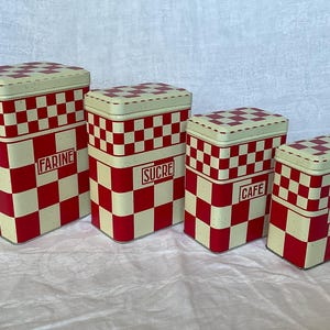 Può includere: Un set di quattro contenitori rettangolari in metallo di dimensioni decrescenti, caratterizzati da un motivo a scacchi rosso e crema. Ogni coperchio e corpo presenta il motivo, con etichette rosse FARINE, SUCRE, CAFE e THE sul davanti.