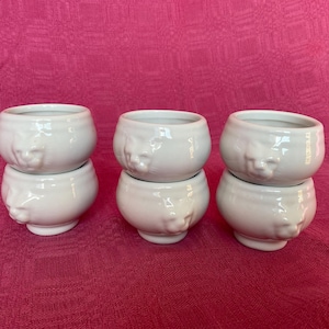 Può includere: Sei tazze in ceramica bianca con un design a forma di viso. Le tazze sono impilate a coppie.