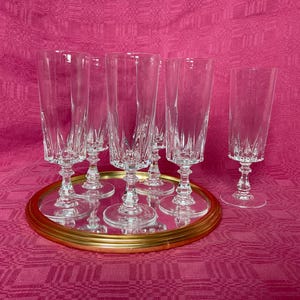 Può includere: Un gruppo di flûte da champagne in cristallo trasparente con intricati disegni incisi. Le flûte sono disposte su un vassoio a specchio con bordo dorato, su uno sfondo rosa acceso. Elegante cristalleria per occasioni speciali.