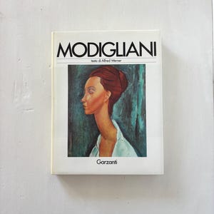 May include: A book titled "MODIGLIANI" with a portrait of a woman on the cover. The portrait features a woman with red hair and a white shirt against a teal and blue background. The publisher's name, Garzanti, is at the bottom.
