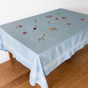 May include: A rectangular light blue tablecloth featuring scattered embroidered floral and fruit motifs in red, green, and yellow. The fabric has a white double-striped border detail along the edges, providing a clean and structured finish to the design.