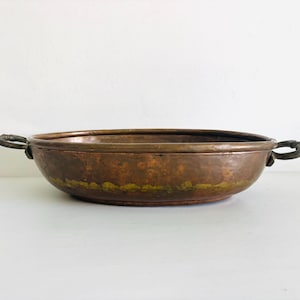 May include: A shallow, oval-shaped copper pan with a hammered texture and a warm, metallic brown finish. The pan features two dark, curved metal handles attached to opposite sides, providing a functional grip for this kitchen vessel.