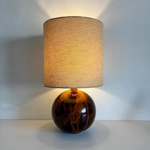 May include: A table lamp featuring a spherical base made of dark brown and amber swirled glass. The lamp is topped with a cylindrical lampshade in a neutral beige fabric, which emits a warm, soft glow when illuminated from within the shade.