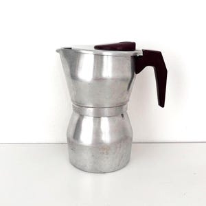 Op de afbeelding: Een zilverkleurige aluminium espressopot met een donkerbruin kunststof handvat en een bijpassende donkerbruine dekselknop. De koffiezetter heeft een klassieke achthoekige vorm met een brede basis, een taps toelopend middengedeelte en een tuit.