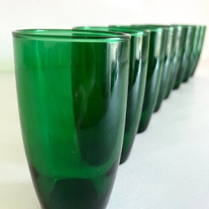 May include: A row of ten green glass tumblers lined up on a white surface. The glasses are all the same size and shape, and they have a slightly rounded bottom.