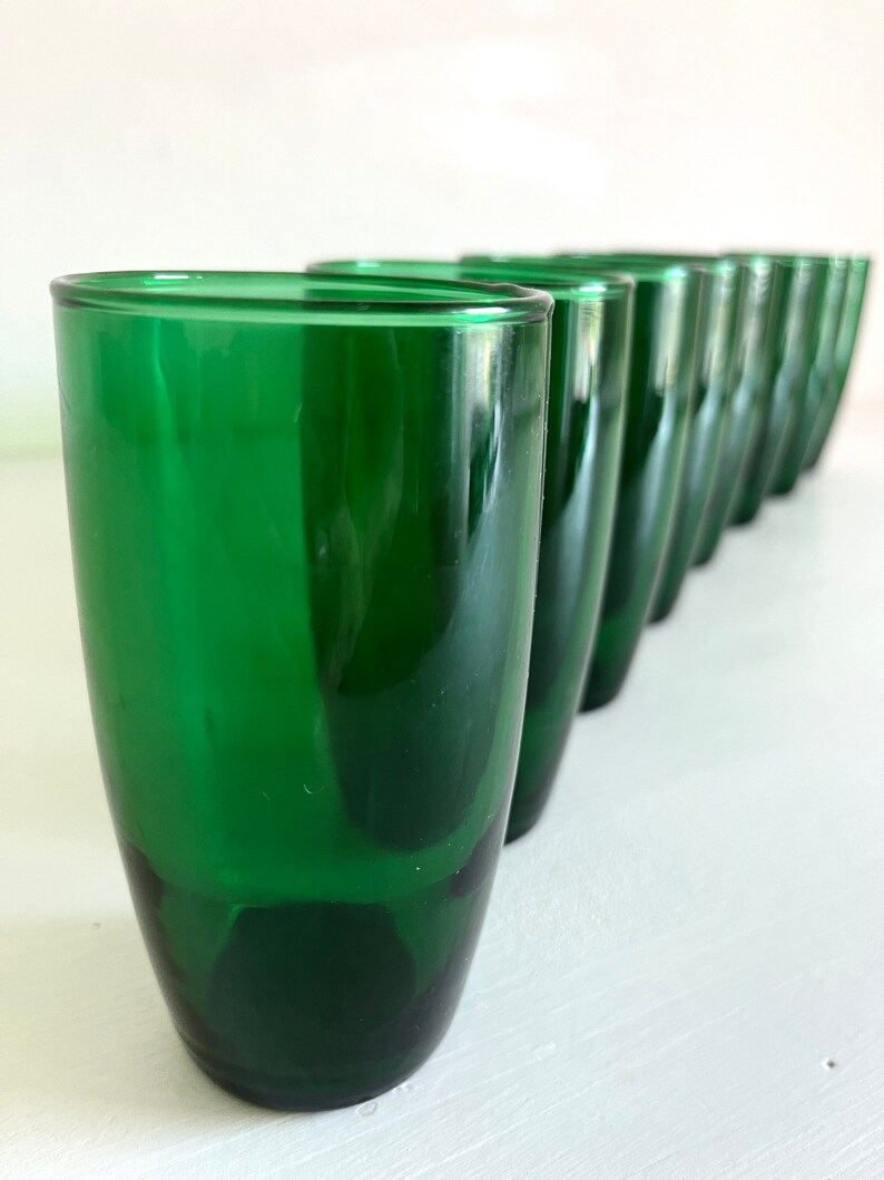 May include: A row of ten green glass tumblers lined up on a white surface. The glasses are all the same size and shape, and they have a slightly rounded bottom.