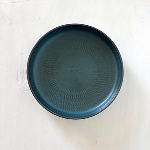 Op de afbeelding: Een teal-kleurige keramische plaat met een donkerblauwe rand. De plaat heeft een getextureerd, geribbeld oppervlak dat vanuit het midden uitstraalt. De plaat is rond en lijkt nieuw.
