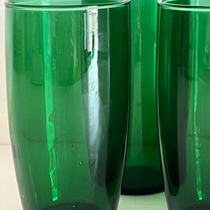 May include: Three tall, green glass tumblers. The glasses are clear and have a smooth, rounded shape.