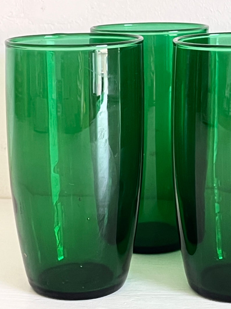 May include: Three tall, green glass tumblers. The glasses are clear and have a smooth, rounded shape.