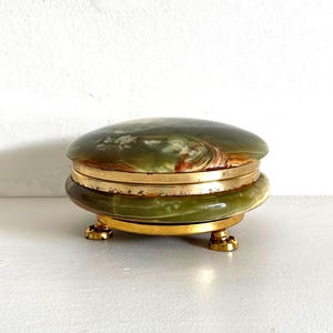 Könnte beinhalten: Eine runde Schmuckdose aus grünem Onyx mit polierter Oberfläche, die natürliche Wirbelmuster in den Farben Dunkelgrün, Creme und Braun aufweist. Die Dose ist von einem goldfarbenen Metallrand eingefasst und steht auf drei kleinen, verzierten goldfarbenen Füßen.