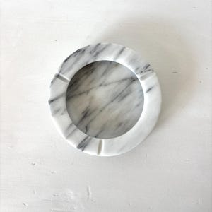 May include: A circular ashtray crafted from white marble, featuring grey veining. The ashtray has a ring design with four small grooves for cigarettes. The interior is smooth and polished.