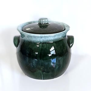 May include: A ceramic jar with a lid, featuring a dark green glaze that transitions to a lighter blue-green at the rim. The jar has two side handles and a knob on the lid. The overall shape is round and bulbous.