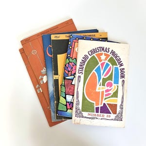 May include: A collection of vintage Christmas program books. The covers feature colorful illustrations and text, including "Standard Christmas Program Book" and "Five Christmas Programs". The books are arranged in a fan shape against a white background.