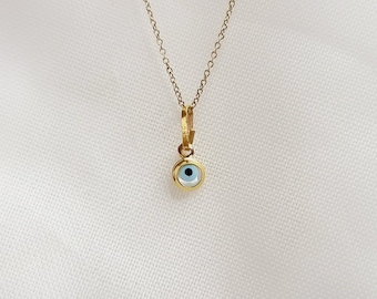 May include: A delicate gold-colored necklace featuring a small pendant with a light blue eye design. The pendant is round, with a gold-colored border. The necklace hangs on a white background.