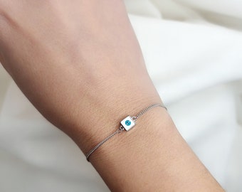 May include: A silver chain bracelet with a small square charm featuring a blue evil eye design.